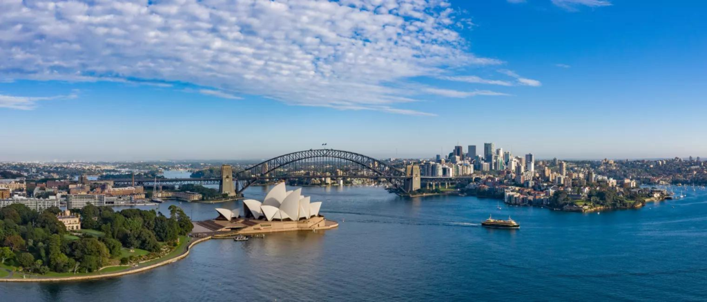 An aerial view of Sydney featuring the city skyline, Sydney Harbour, Opera House, and Harbour Bridge.
