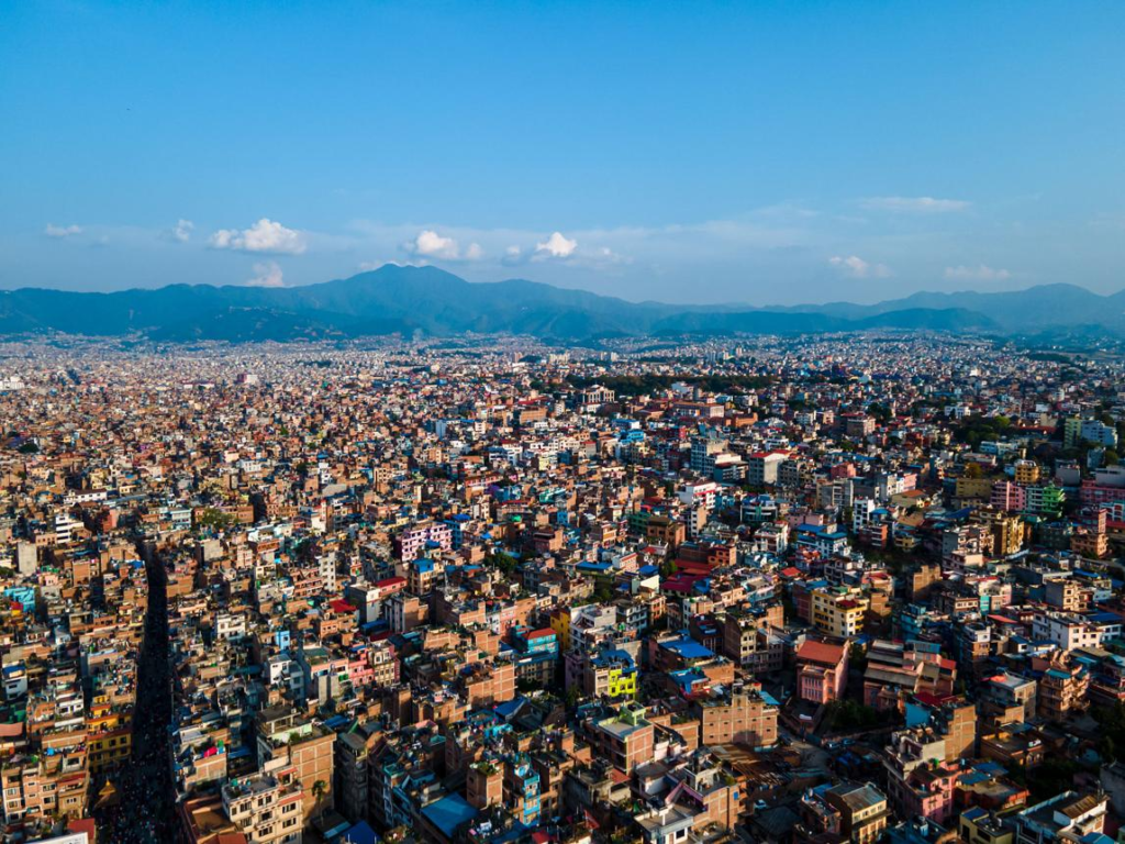 A panoramic aerial view of Kathmandu showing the cityscape, surrounding hills, and historic buildings with the Himalayas in the distance.